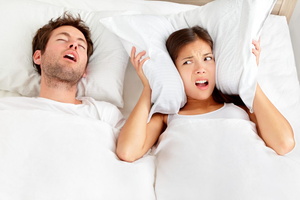 Sleep Apnea. Snoring man. Couple in bed, man snoring and woman can not sleep, covering ears with pillow for snore noise. Young interracial couple, Asian woman, Caucasian man sleeping in bed at home.